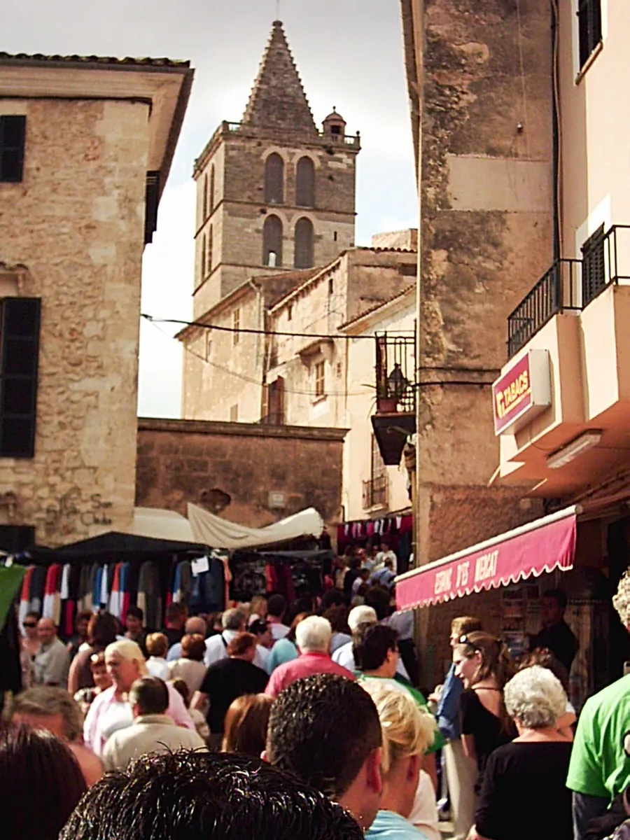 Menschen zwischen Marktständen in einer Gasse von Sineu, dahinter der Kirchturm